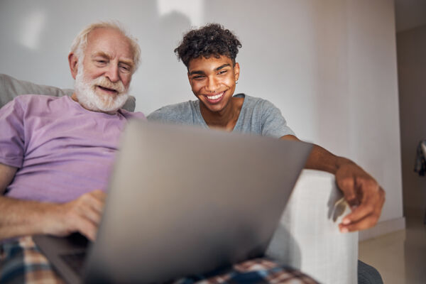 Photo shows a young man with brown skin and dark hair next to an old man with white skin, and white hair spending time looking at something on the laptop.